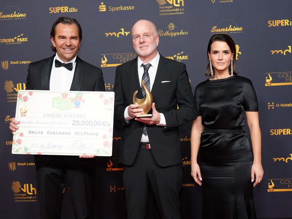 Robert Dahl, Peter Nitschke und Yara Hoffmann stehen vor einer Fotowand mit Trophäe und großem Spendenscheck in der Hand.